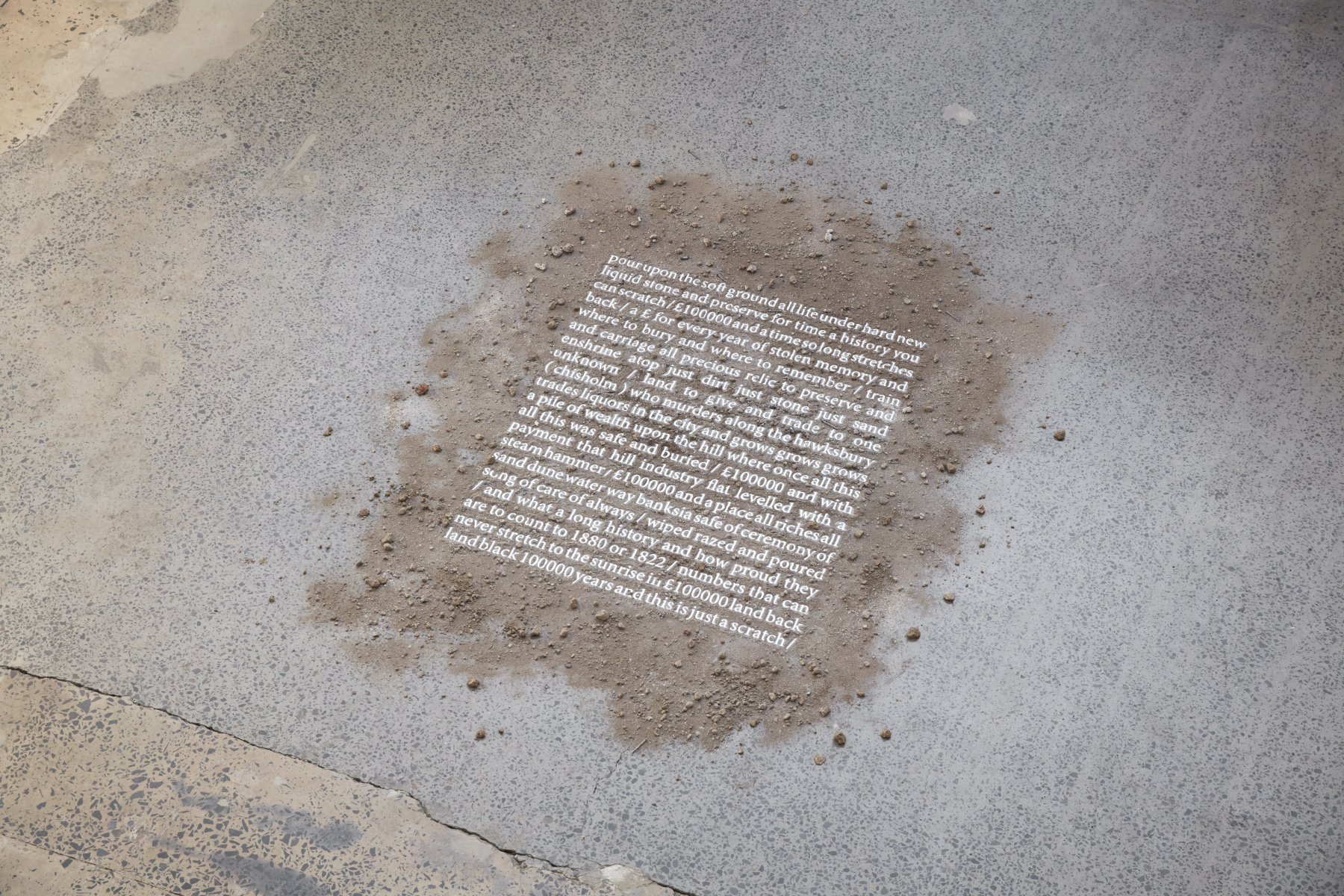 A poem written in white text is revealed through dirt laid upon a concrete ground.
